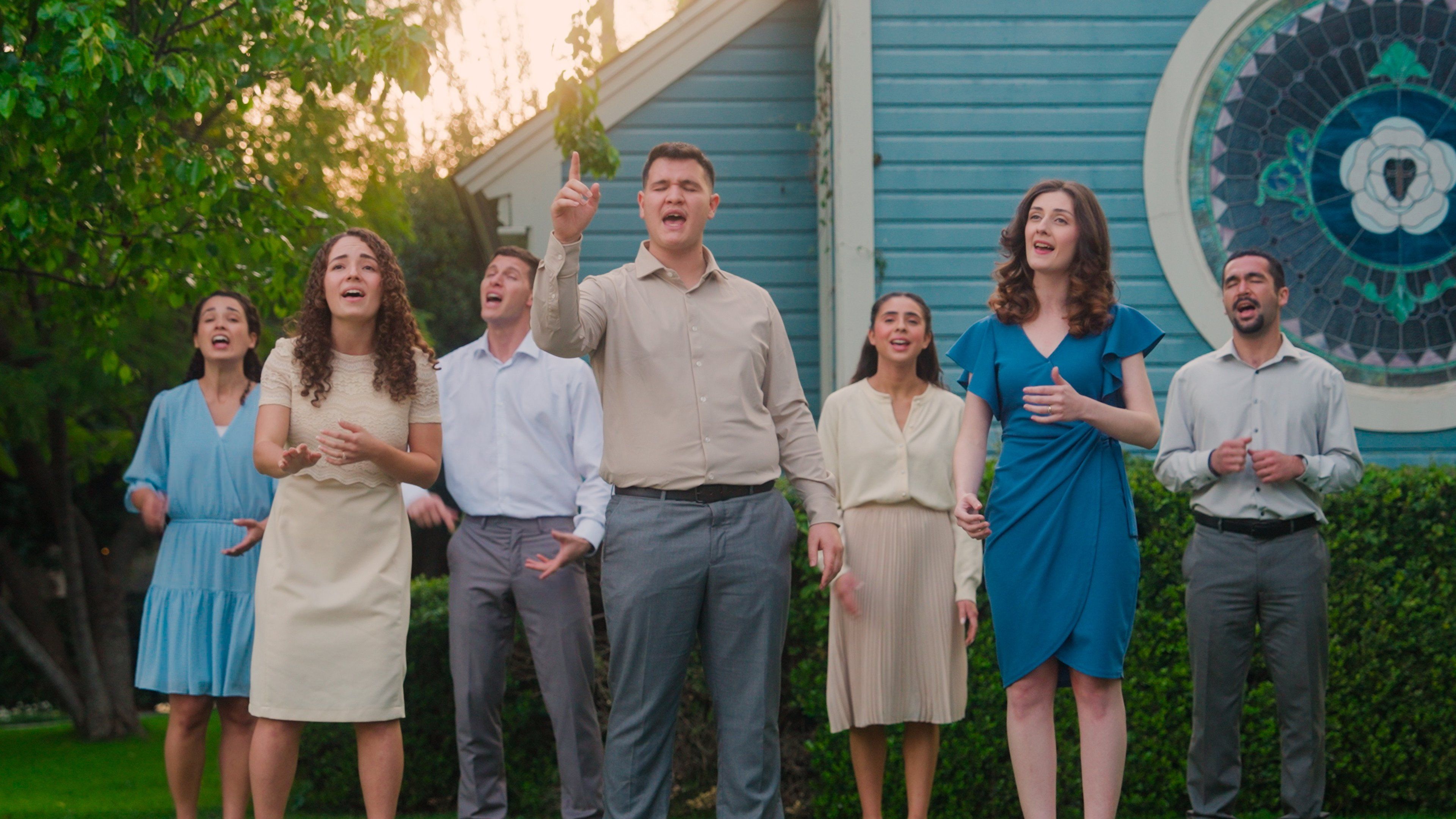 The seven members of Abrigo Vocal singing outdoors in front of a blue church at golden hour