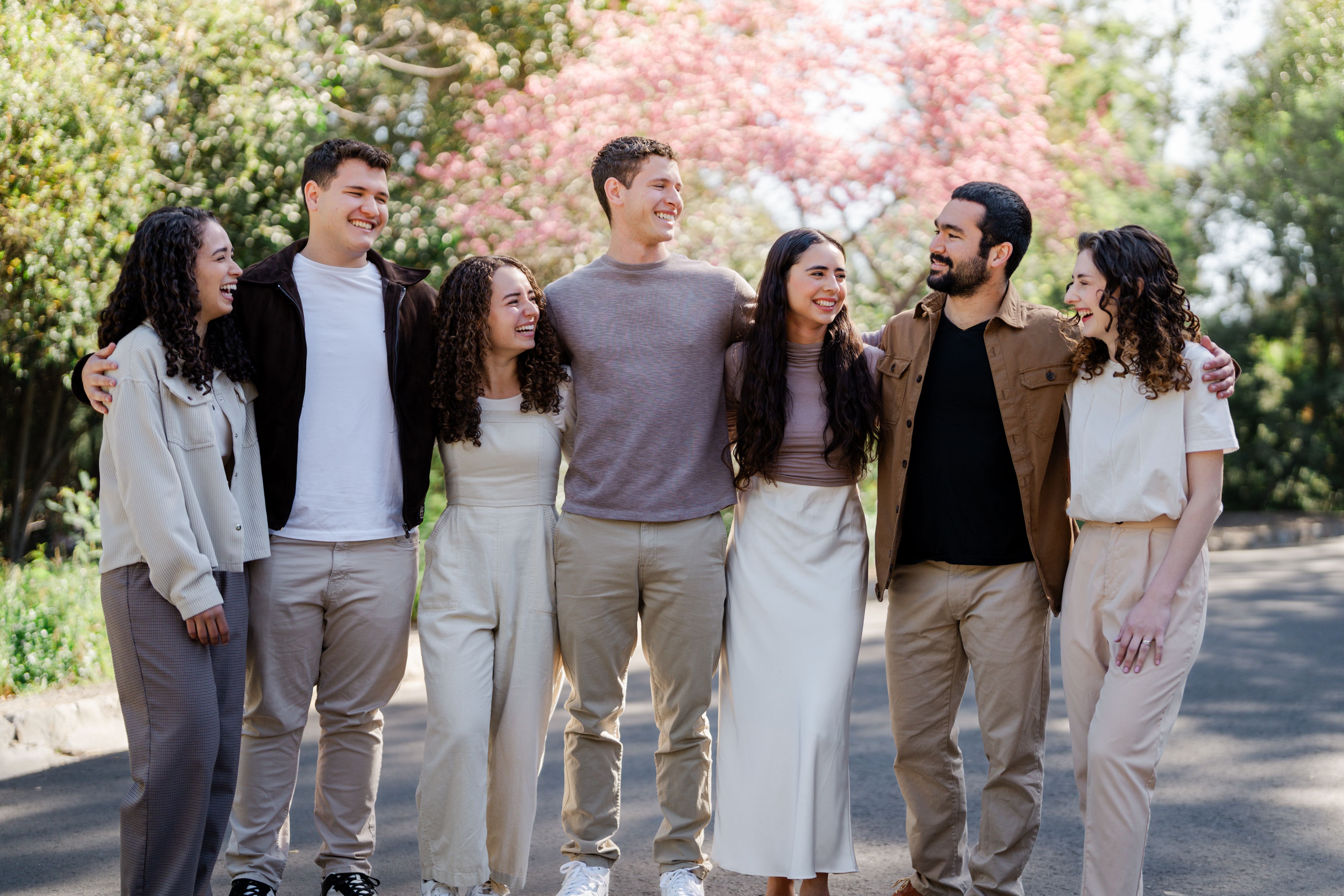 The seven members of Abrigo Vocal standing arm in arm and laughing together outdoors, with trees and pink blossoms behind them